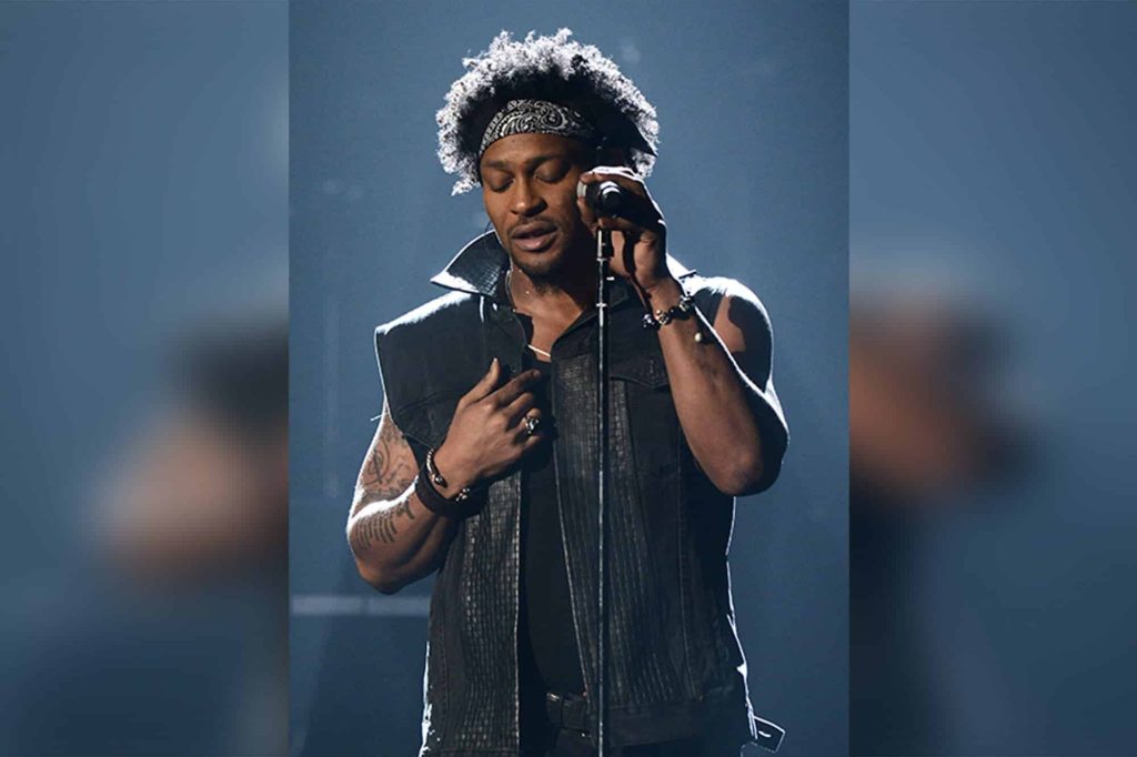 "Tribute photo as D Angelo dies at 51 D’Angelo onstage singing into a mic, wide-brim hat, moody lights (2012 Bonnaroo)"